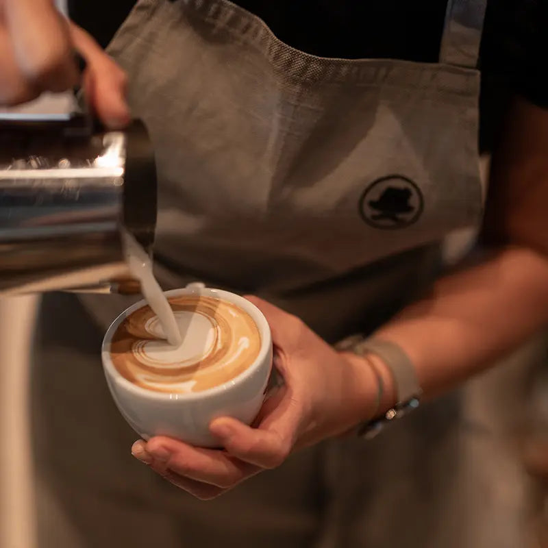Barista mit Kuntrawant-Schürze gießt Milchschaum für Latte Art in Tasse
